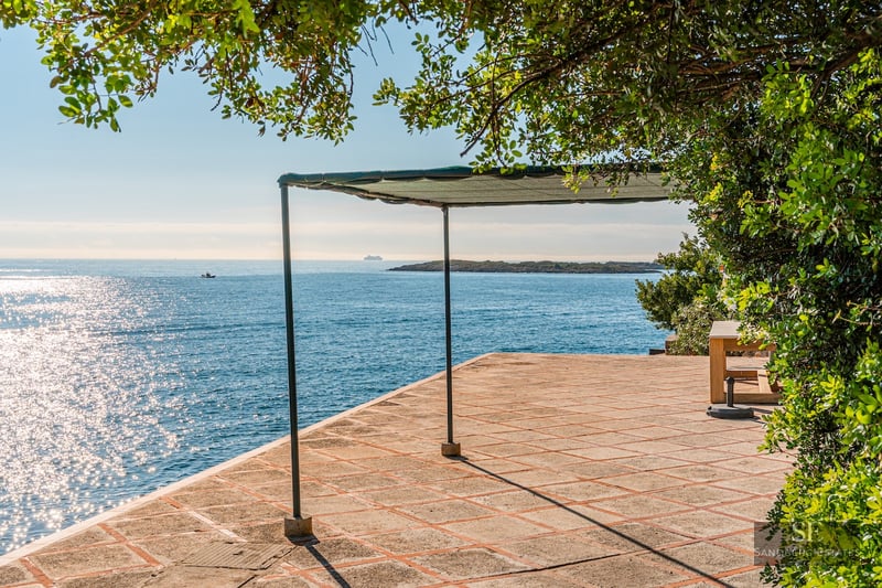A sun-drenched stone terrace with a shade structure overlooking the sparkling blue ocean and a distant island.
