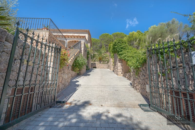 Open wrought iron gates lead up a paved driveway flanked by stone walls and lush greenery under a clear blue sky.