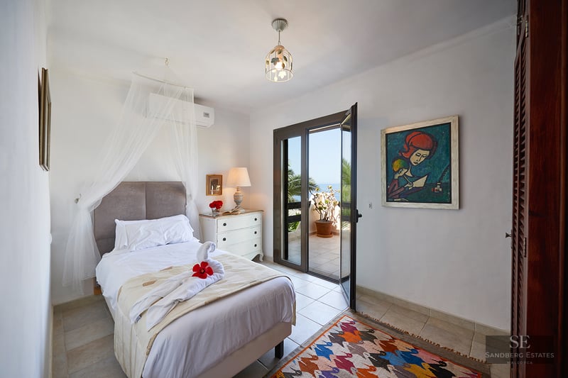 A white bedroom with a mosquito net over the bed, a colorful rug, and open glass doors leading to a sunny terrace.
