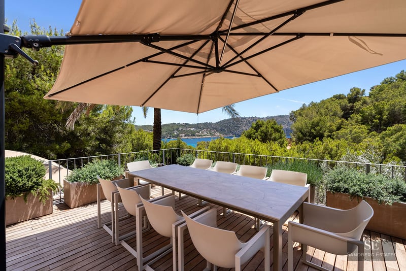 Outdoor wooden terrace with a dining table and chairs under a large umbrella overlooking a sea bay.