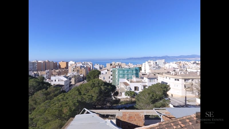 High-angle view of a coastal city with white buildings, green pine trees, and the blue Mediterranean sea in the background.