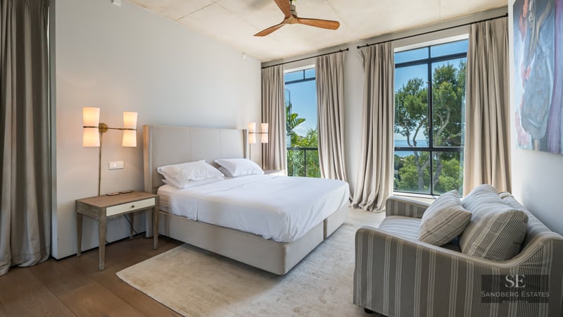 Bright master bedroom with a large bed, striped sofa, and floor-to-ceiling windows overlooking lush trees and the sea.