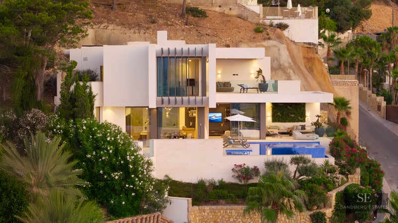 Modern white multi-level villa with a swimming pool, terraces, and large glass windows built into a rocky hillside.