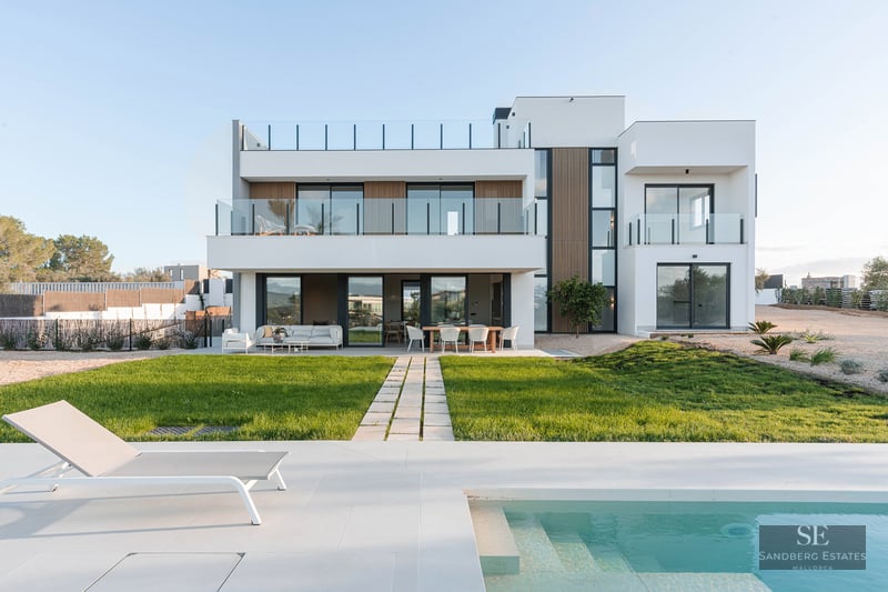 Modern white two-story villa with a turquoise swimming pool, sun lounger, and a green lawn under a clear sky.