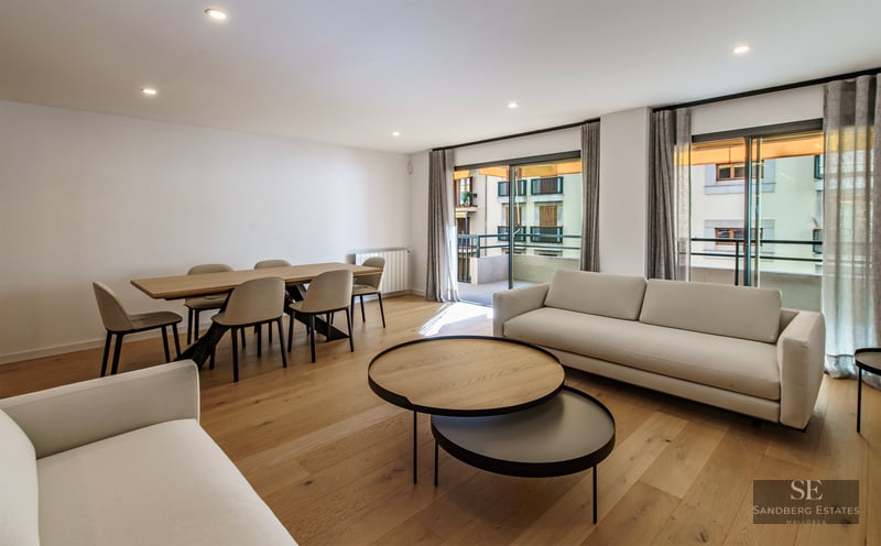 Salon moderne avec canapés blancs, tables basses en bois, coin repas et grandes baies vitrées ouvrant sur un balcon.