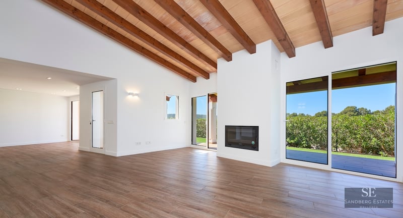 Bright, empty living room featuring exposed wooden ceiling beams, a built-in fireplace, and large glass doors to a garden.