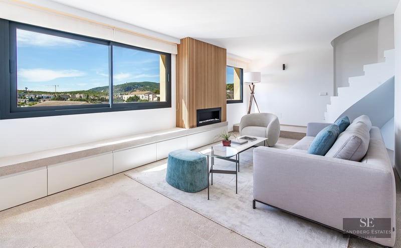 Bright living room featuring a grey sofa, teal ottoman, wood slat fireplace wall, and large window overlooking hills.