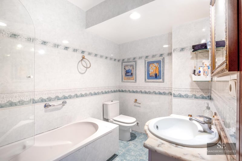 Bright bathroom featuring tiled walls, a bathtub with glass partition, white toilet, and marble sink countertop.