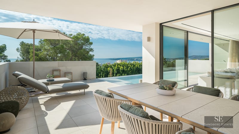 Modern terrace with outdoor dining, lounge chairs, and an infinity pool overlooking the Mediterranean sea and trees.