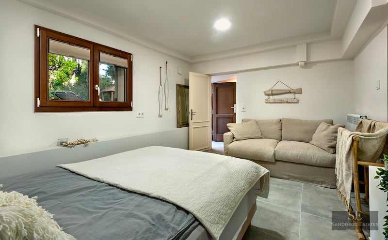 A bright bedroom featuring a grey bed, a beige sofa, a wooden window, and nautical-themed wall decor.