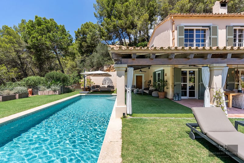 Rectangular turquoise pool next to a Mediterranean villa with a covered terrace and green shutters under a blue sky.