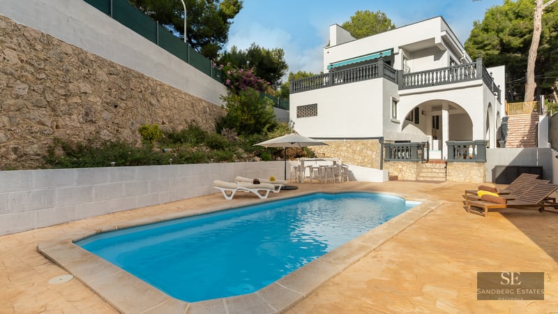 Piscine rectangulaire à l'eau turquoise entourée d'une terrasse en pierre, de chaises longues et d'une villa blanche.