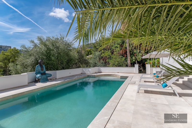 Turquoise pool with white stone tiling, lounge chairs, and a Buddha statue under palm trees.