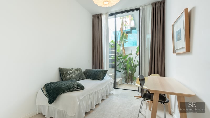 Bright bedroom with a single bed, wooden desk, and glass door opening to a small outdoor garden with plants.