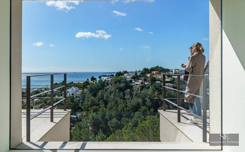 Two people on a modern terrace overlook a lush green coastal landscape meeting the blue Mediterranean Sea.
