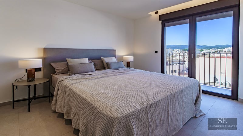 Modern bedroom featuring a large bed with grey bedding, stylish lamps, and a glass door overlooking a city and mountains.