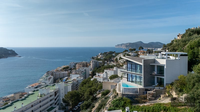 Modern white villa on a green hillside with a small infinity pool overlooking a coastal town and the blue sea.