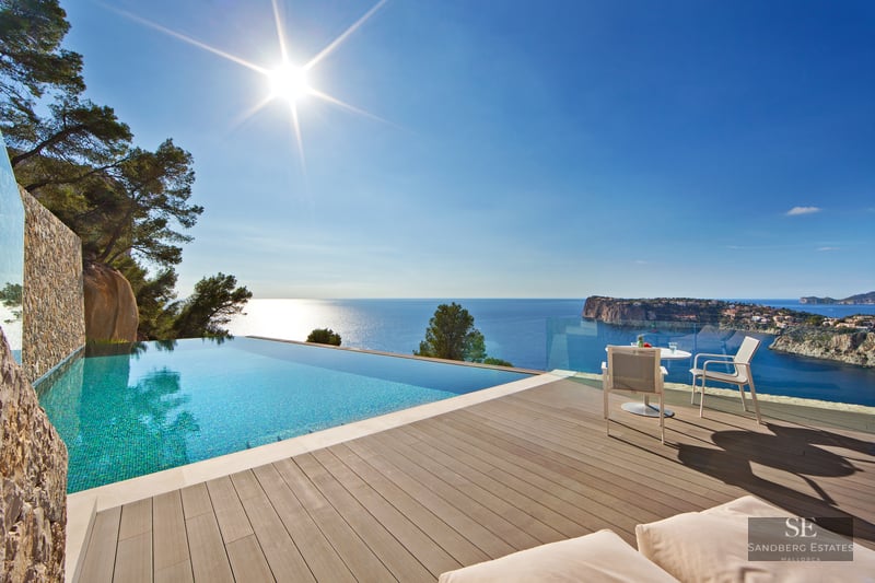 Infinity pool with turquoise water on a wooden deck overlooking a blue sea and rugged coastline under a bright sun.