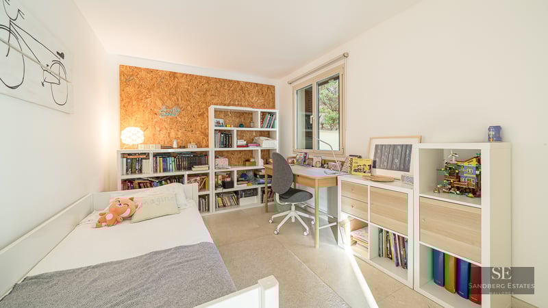 Child's bedroom featuring a white bed, wooden desk, and extensive shelving filled with books under a bright window.