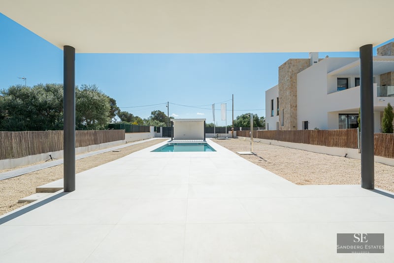 View from a covered porch toward a rectangular pool surrounded by white tiles and gravel in a modern villa setting.