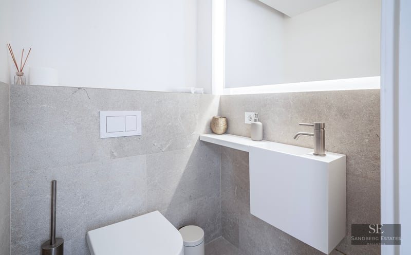 Salle de bain minimaliste avec WC blanc, petit lavabo suspendu et grand miroir rétroéclairé sur murs en pierre grise.