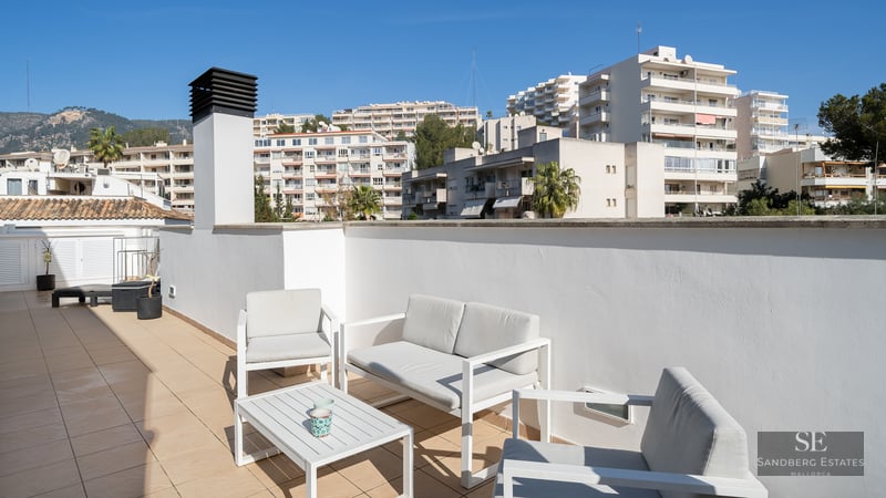 Sunlit rooftop terrace with white modern furniture, tiled floor, and views of city buildings and mountains.