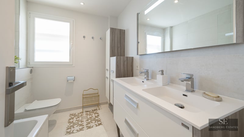Bright modern bathroom with a double white vanity, large mirror, wall-hung toilet, and natural window light.