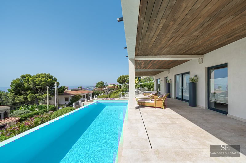A bright terrace with a turquoise infinity pool, wicker loungers, and a wooden ceiling overlooking a coastal landscape.