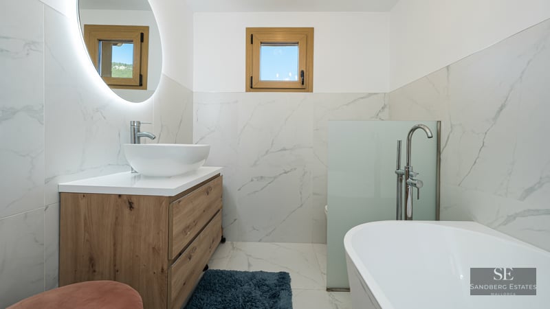 Modern bathroom with marble walls, wood vanity, vessel sink, circular backlit mirror, and a white bathtub.