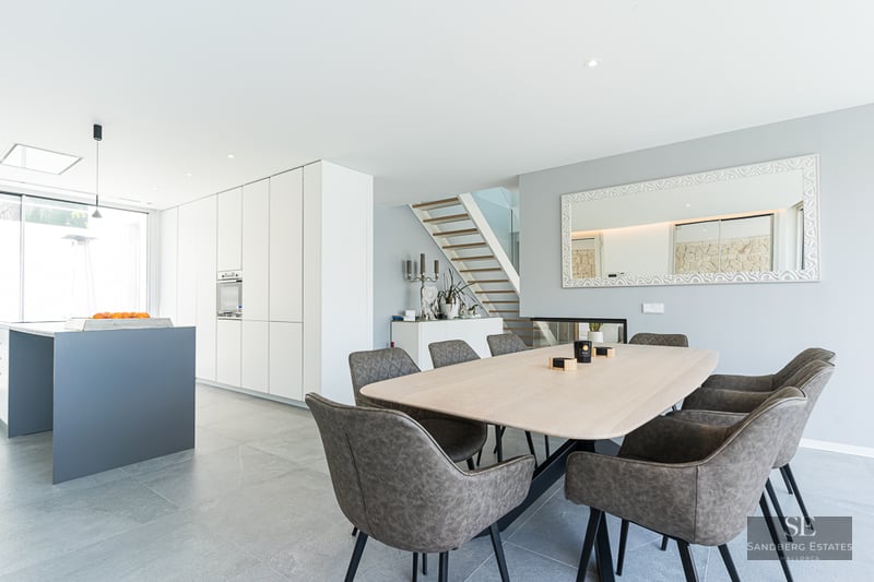 Bright dining area featuring a light wood table, grey chairs, and a minimalist white kitchen in the background.