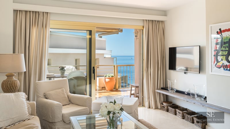 Elegant living room with beige armchair and glass table opening to a terrace with a panoramic Mediterranean sea view.