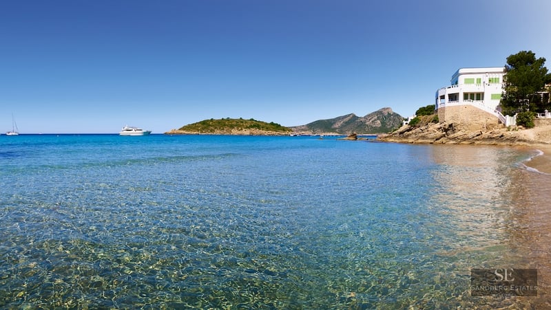 View of a crystal clear turquoise bay with a white villa on a cliff and mountains in the distance.