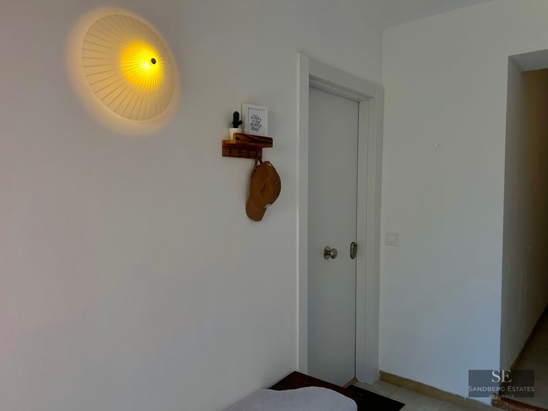 A white hallway featuring a round pleated wall lamp, a small wooden shelf with a hat, and a white door.