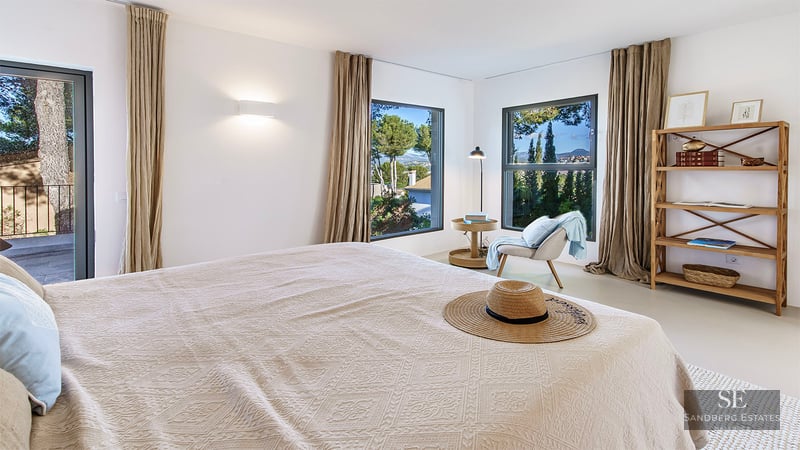 Modern bedroom with white walls, linen curtains, wooden furniture, and windows overlooking a lush green landscape.