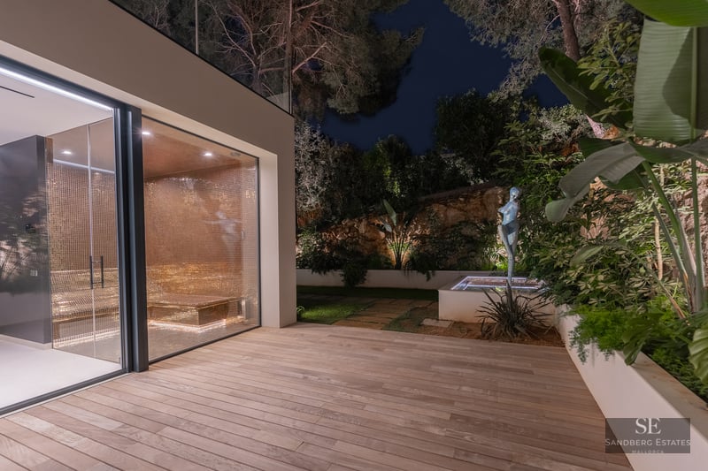 Glass-enclosed spa with gold mosaic tiles next to a wooden deck and illuminated garden with a sculpture at night.