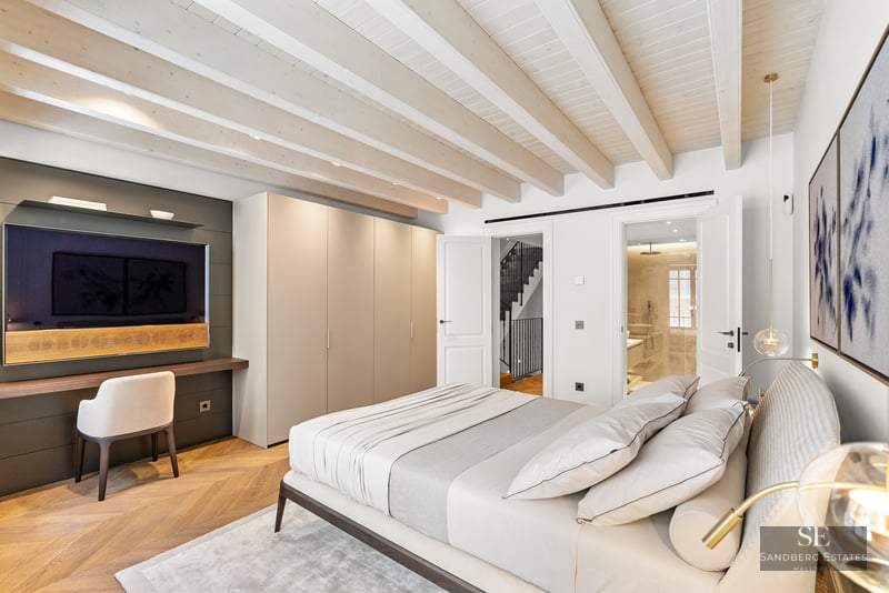 Luxury bedroom with white beamed ceiling, ensuite bathroom, built-in wardrobes, and wall-mounted TV over a desk.