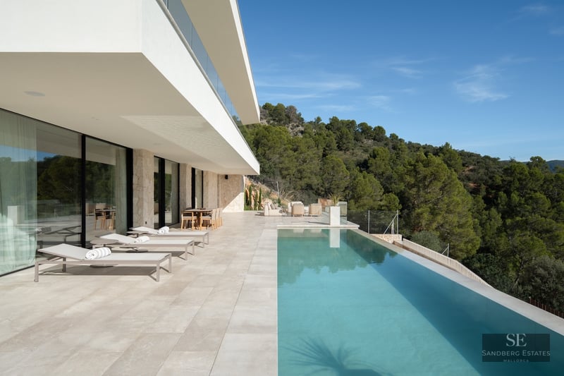 A spacious white stone terrace featuring an infinity pool, sun loungers, and views of a pine forest under a blue sky.