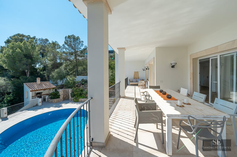 Terrasse couverte blanche avec grande table à manger et vue sur une piscine bleue et de la verdure.