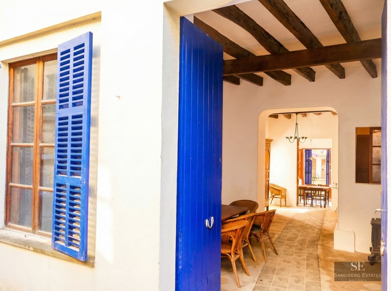 White exterior wall with bright blue shutters and open door leading to a rustic interior with wooden beams.