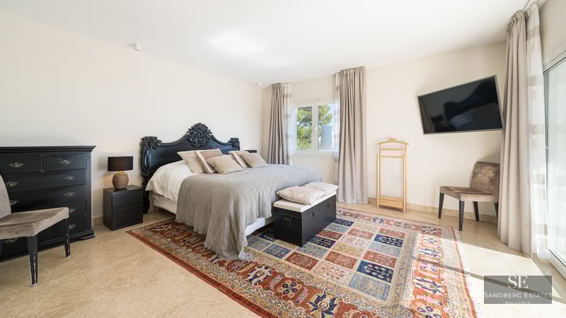 A bright master bedroom with an ornate black headboard, a colorful patterned rug, and travertine tile floors.