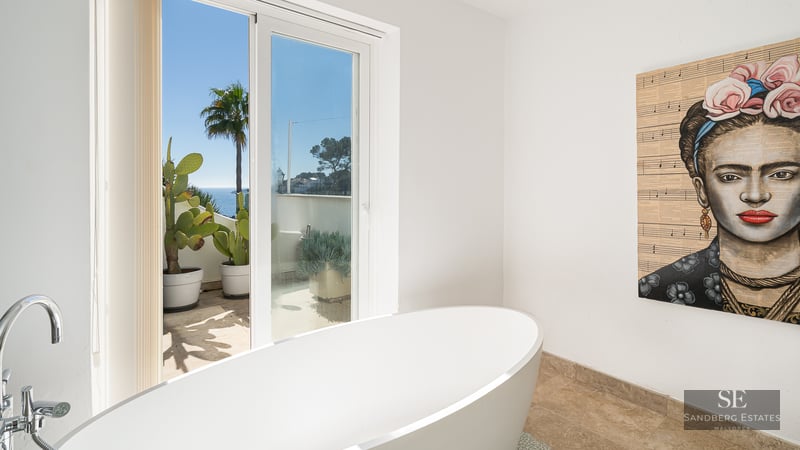 Modern bathroom featuring a white freestanding tub, Frida Kahlo portrait, and glass doors opening to a sea-view terrace.