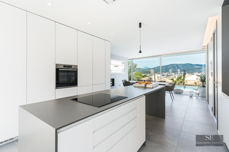 Bright white kitchen with a grey island, induction hob, and large windows overlooking mountains and a pool.