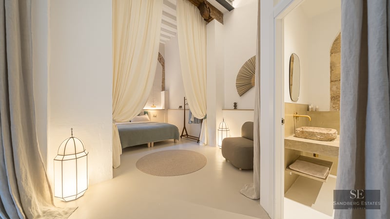 A serene master bedroom with flowing cream drapes, lantern lamps, and a glimpse into a stone-accented ensuite bathroom.
