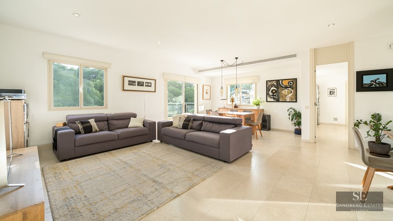 Spacious bright living room with two grey sofas, light tile flooring, and dining area in the background with large windows.