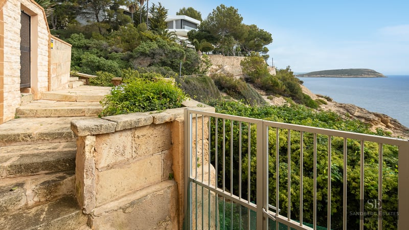 Stone steps and walls leading to a metal gate overlooking the Mediterranean sea and a distant island.