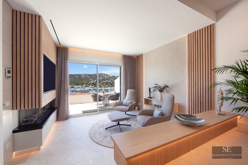 Modern lounge with wood slats, designer chairs, and a sliding glass door revealing a stunning Mediterranean harbor view.