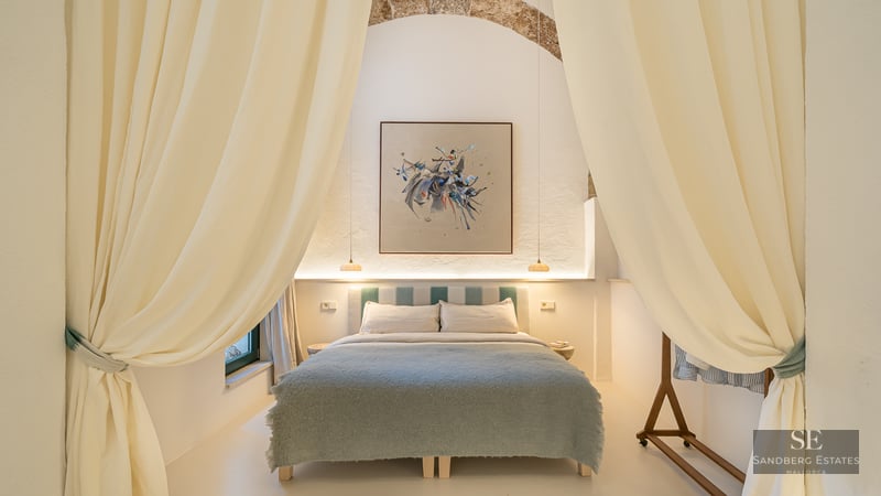 Luxury bedroom framed by cream curtains, featuring a bed with a blue throw, abstract art, and a rustic stone arch.