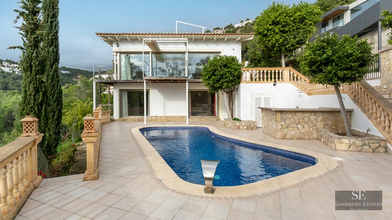 Blue swimming pool on a tiled terrace in front of a white Mediterranean villa with cypress trees.