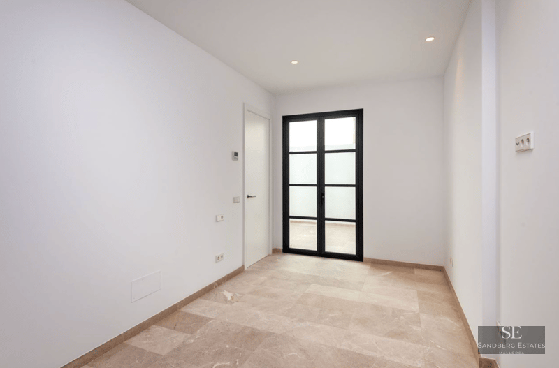Empty room with white walls, beige stone floors, and black-framed glass doors leading outside.