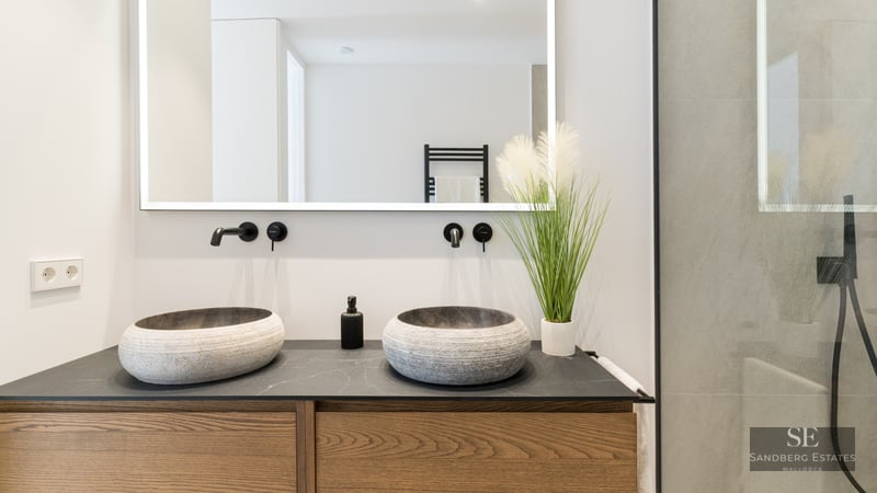 Modern bathroom vanity featuring two stone vessel sinks, black wall-mounted faucets, a backlit mirror, and wooden cabinets.